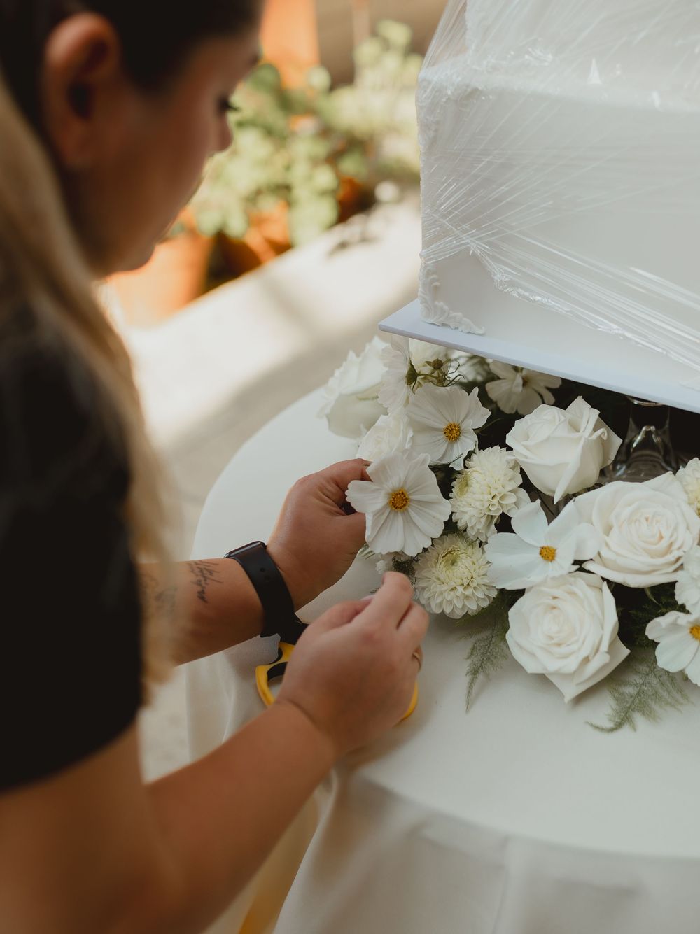 A woman arranges white roses and daisies on a tiered white wedding cake.