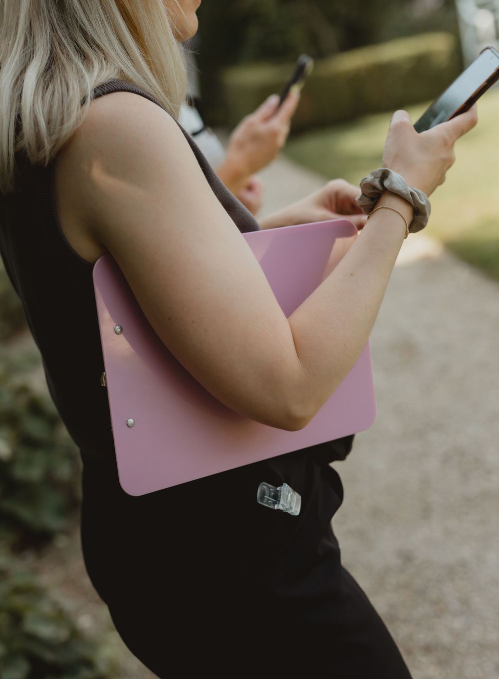 Woman in black dress holding pink clipboard and smartphone outdoors in a garden setting.