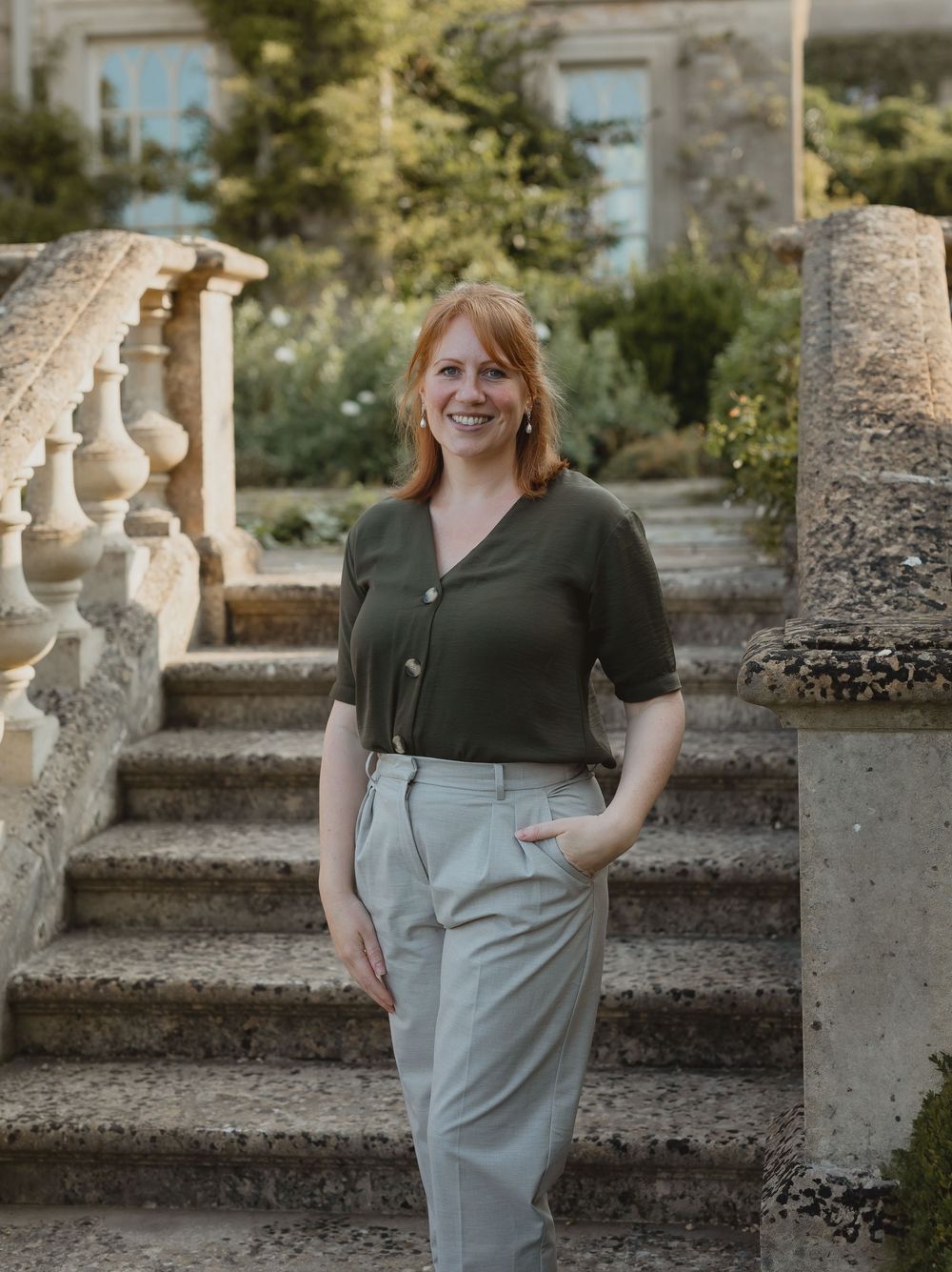 Smiling redhead woman in olive green top and grey trousers standing on stone steps in a garden.