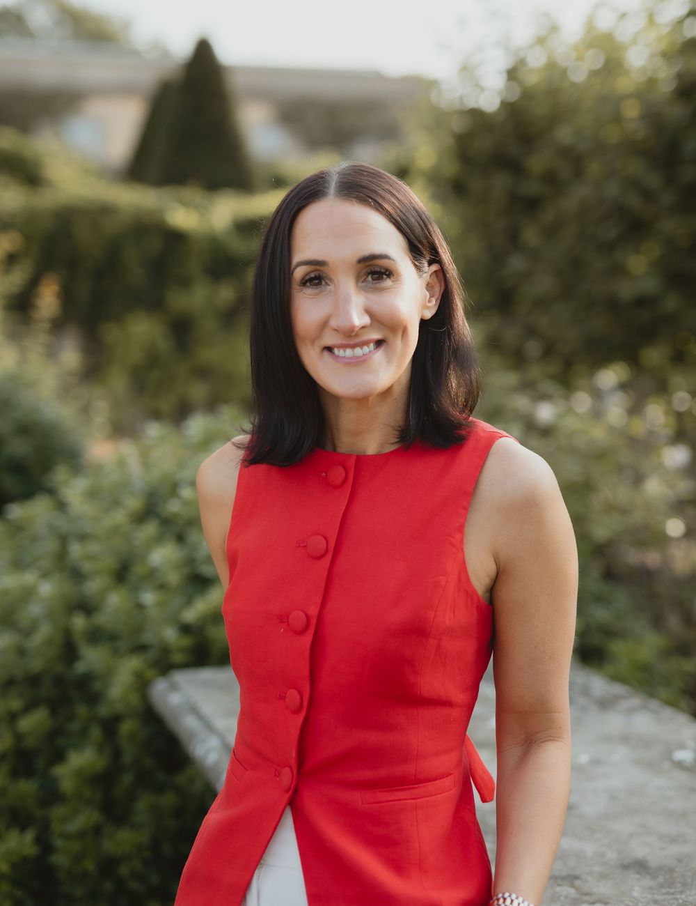 Smiling brunette woman in red sleeveless dress standing in garden with green shrubs in background.