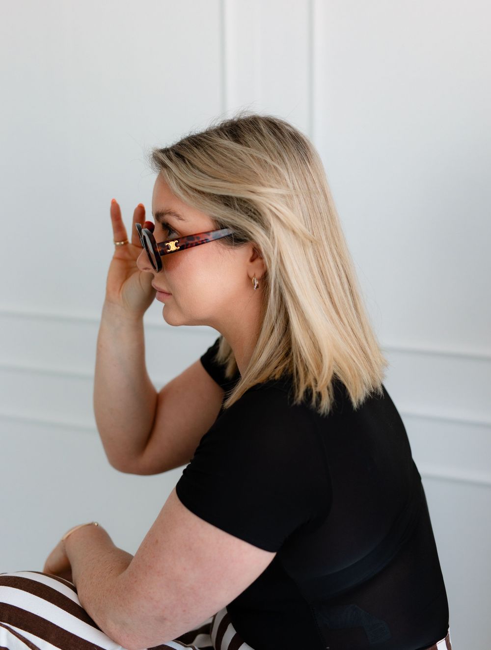 Side profile of someone wearing sunglasses and a black t-shirt posing against a white wall background.