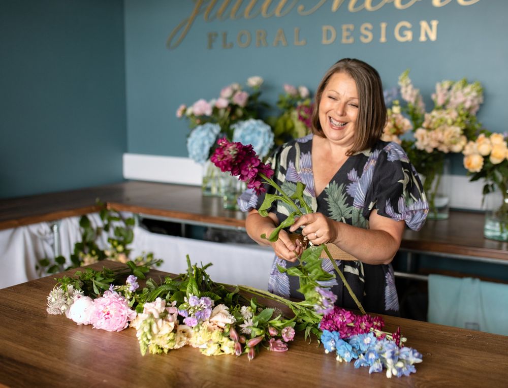 A floral designer works at a wooden table with various colorful blooms and greenery spread out for arrangement.