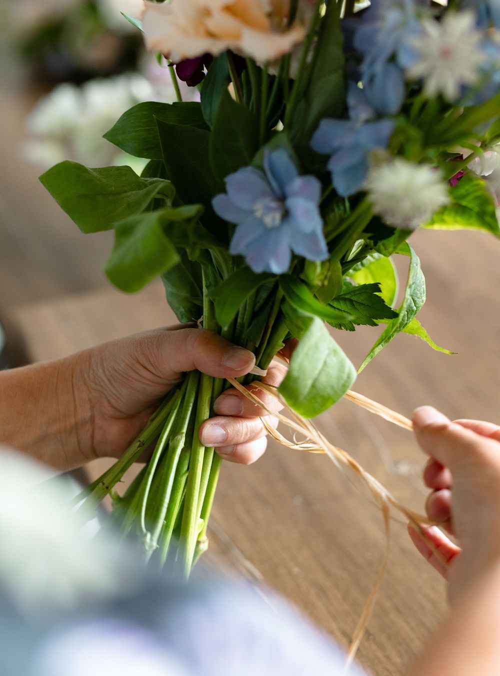 Close-up of hands working with delicate blue and white flowers and greenery on a wooden surface.