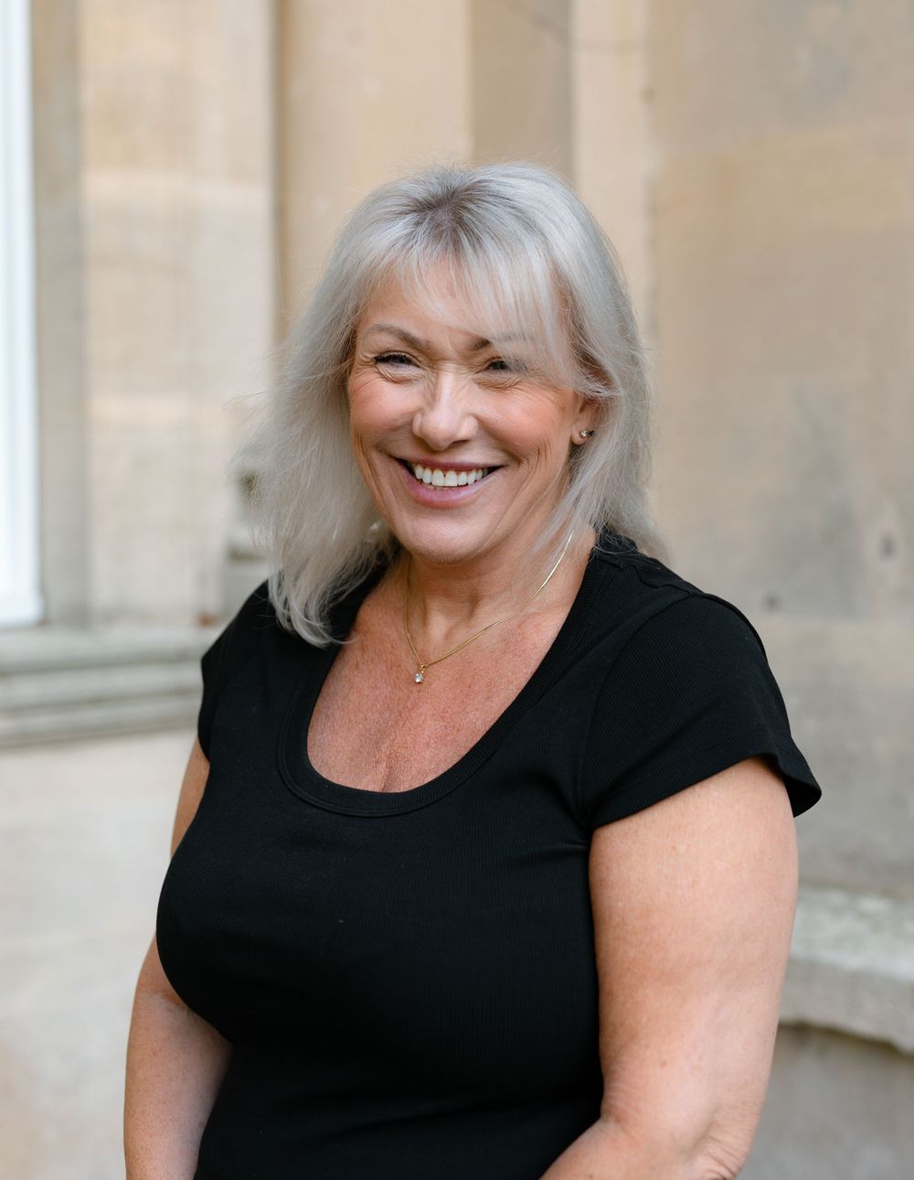 Woman wearing a black top smiles warmly while standing against a light beige wall.
