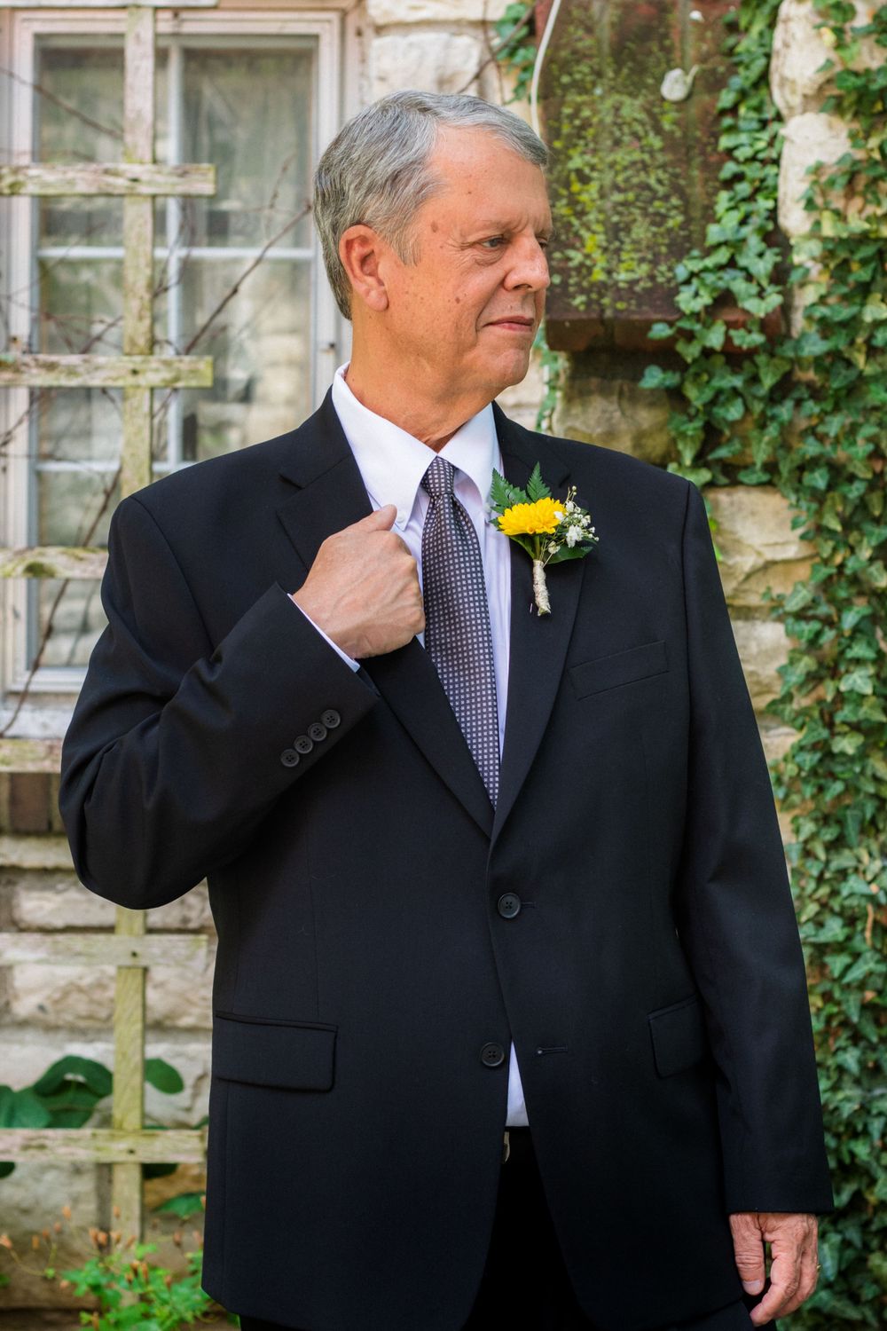Groom in dark suit adjusts his tie against stone and ivy backdrop.