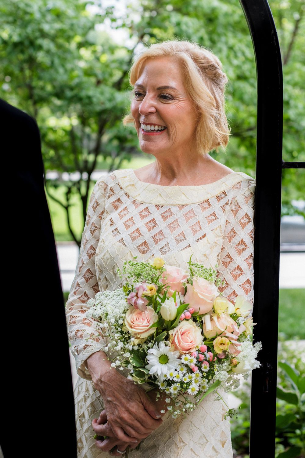 Bride in lace dress holds a delicate bouquet of pink roses and baby's breath.