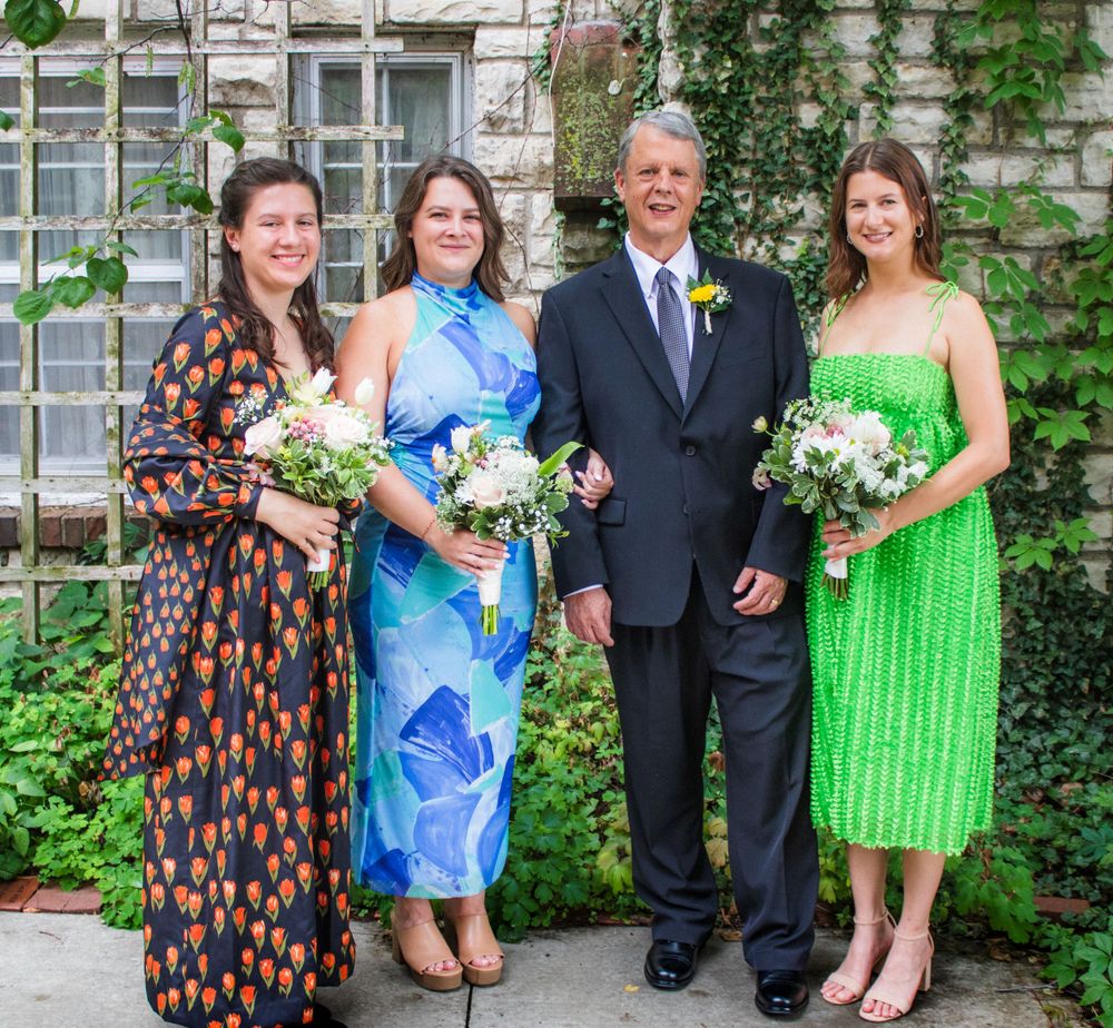 Four people in formal attire pose together outdoors with bouquets against a vine-covered wall.