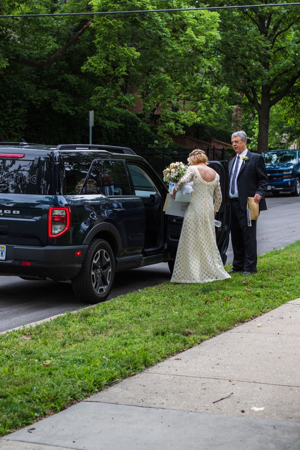 A couple stands next to a black SUV on a residential street during a wedding celebration.
