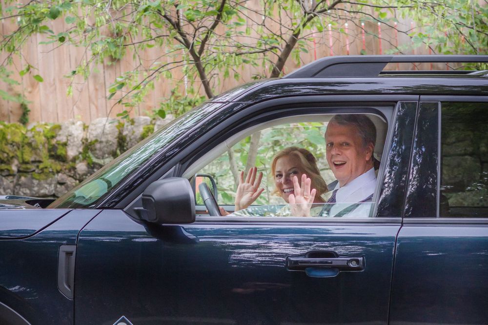 Two people wave excitedly from inside a black car.