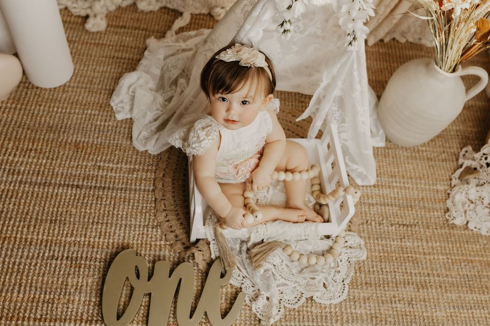 Baby girl in lace outfit sits by a wooden 'one' sign for a boho first birthday cake smash photo session.