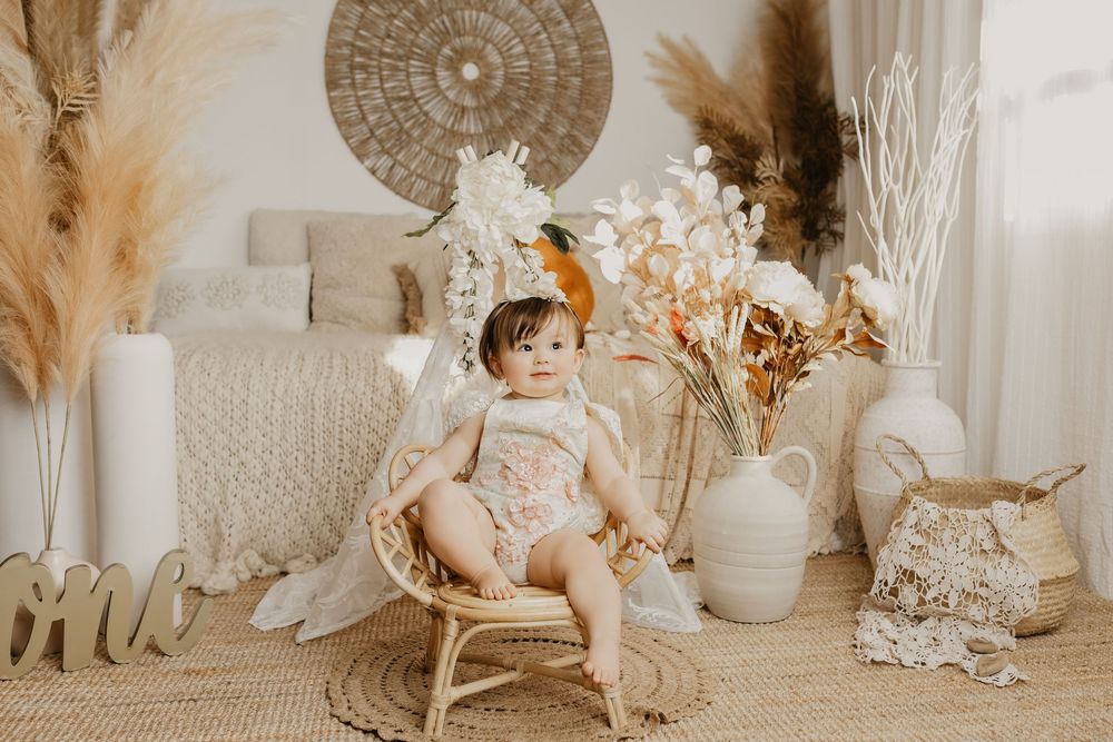 Baby girl in boho romper sits in rattan chair surrounded by pampas grass and white floral decor for first birthday.