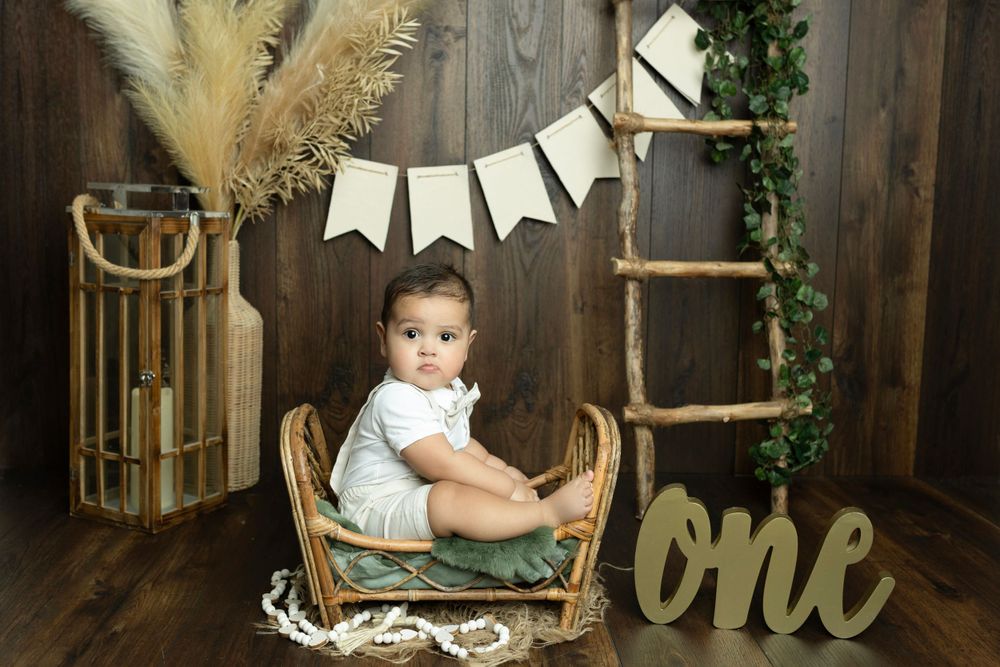 Rustic first birthday setup with wooden backdrop, banner, and wicker chair.