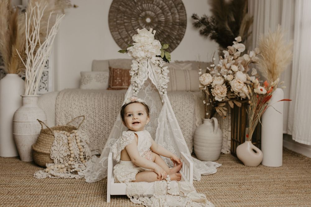 Boho-styled nursery scene with white lace, neutral decor, dried flowers and vintage elements arranged on beige carpet.