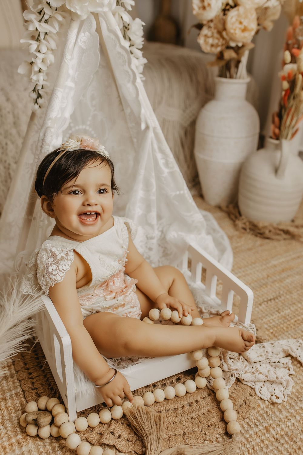Smiling baby girl in white lace romper sits on tiny white bed with boho teepee, wooden beads, and floral vases backdrop.