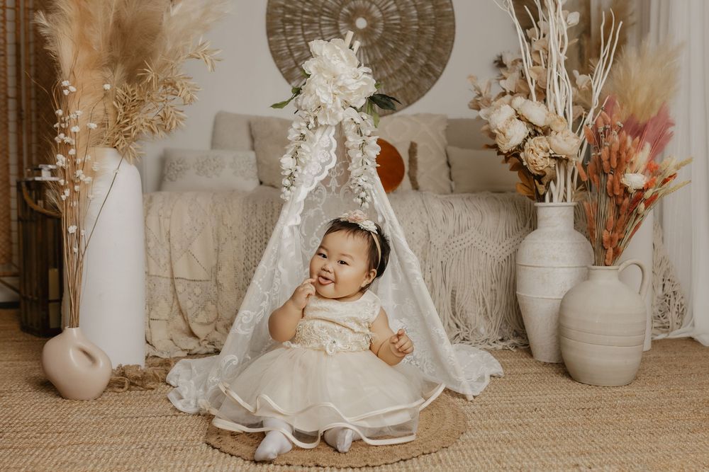 Baby girl in white lace dress sits smiling under a floral teepee in a boho-styled photo studio setting.