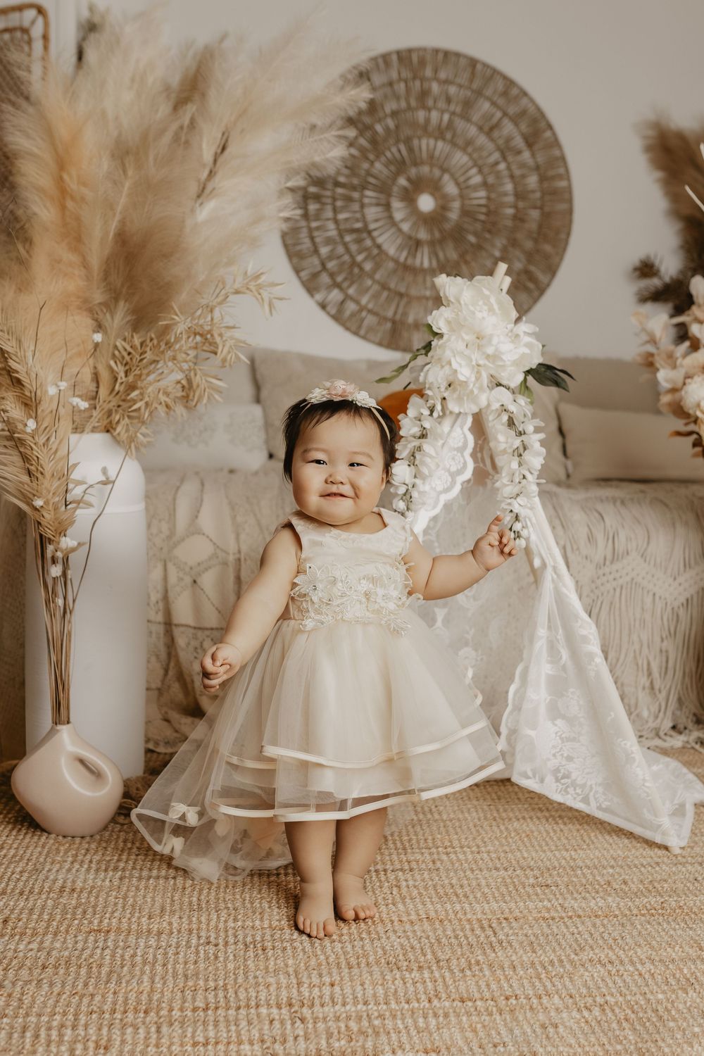 A smiling baby girl in a cream tulle dress holds a white floral hoop, standing barefoot in a boho-styled room.