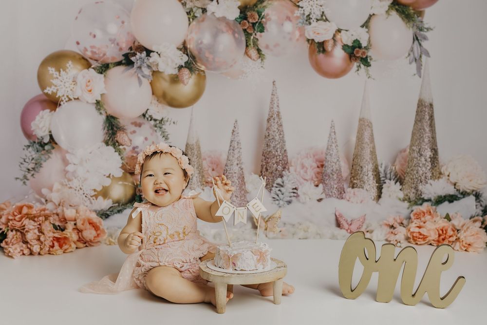 Baby girl in pink lace outfit smiles at first birthday cake smash with floral balloon backdrop and gold ONE sign.
