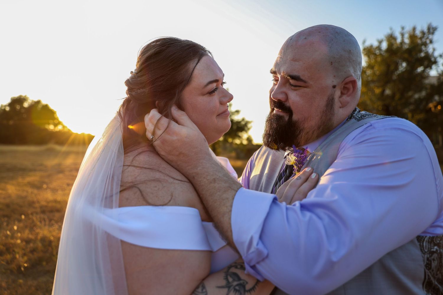 A couple shares an intimate moment at sunset in a field while wearing wedding attire.