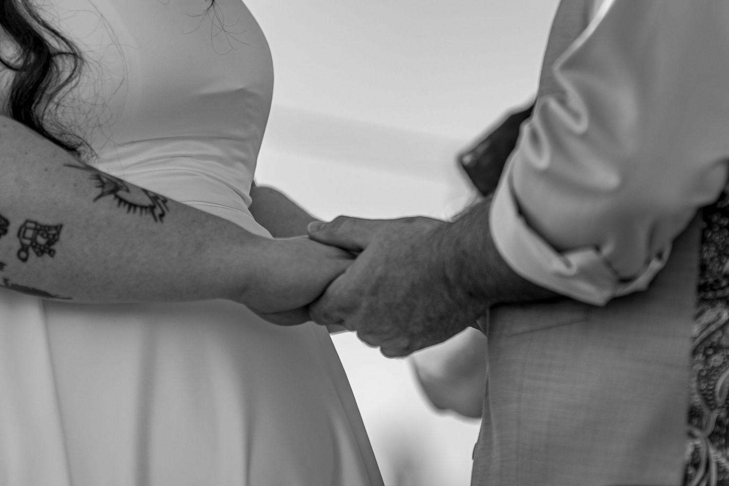 Black and white photo of a couple holding hands at their wedding ceremony, bride with tattoo visible.