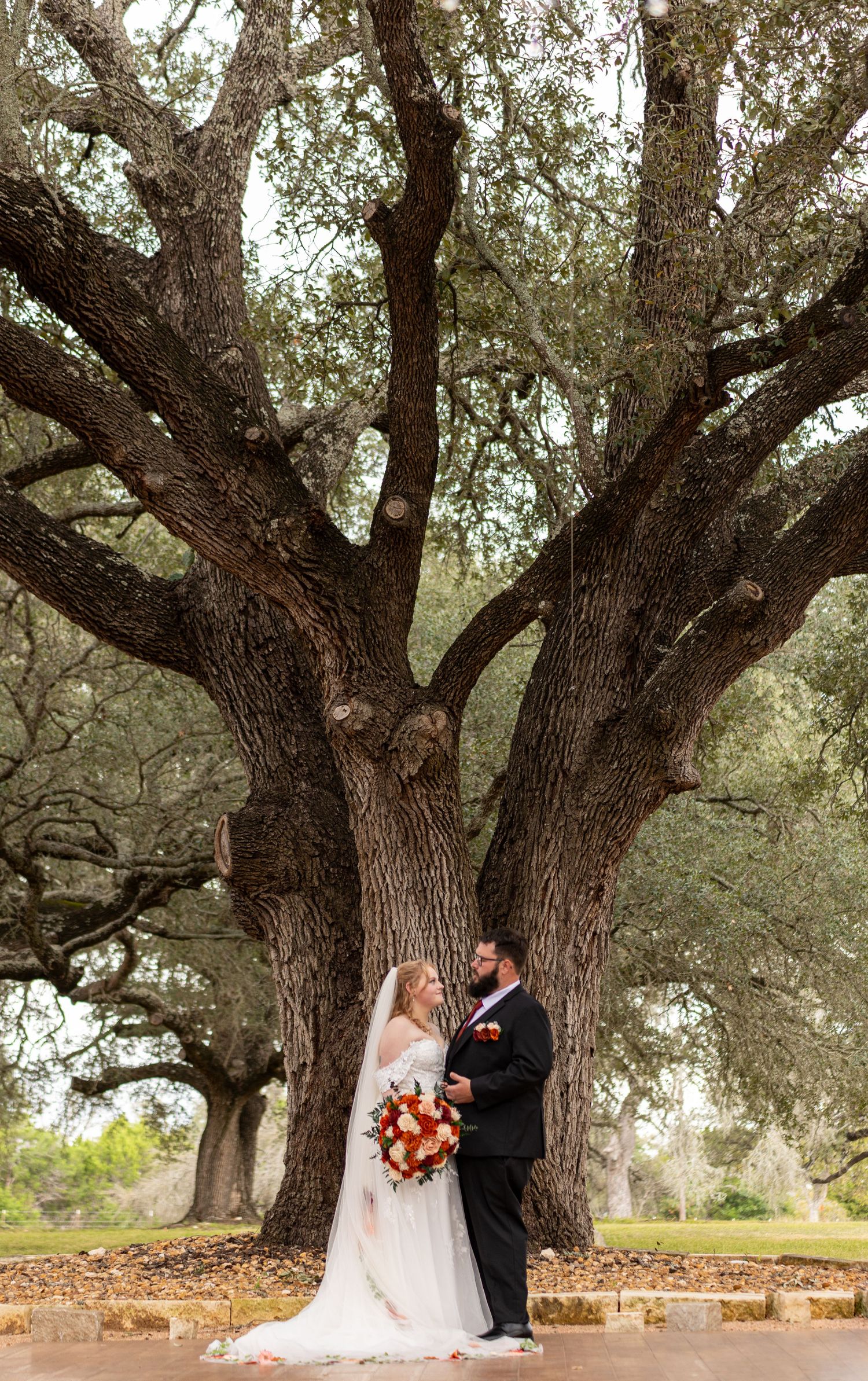 Bride and groom standing together under a massive oak tree, bride holding a red bouquet in a white gown.