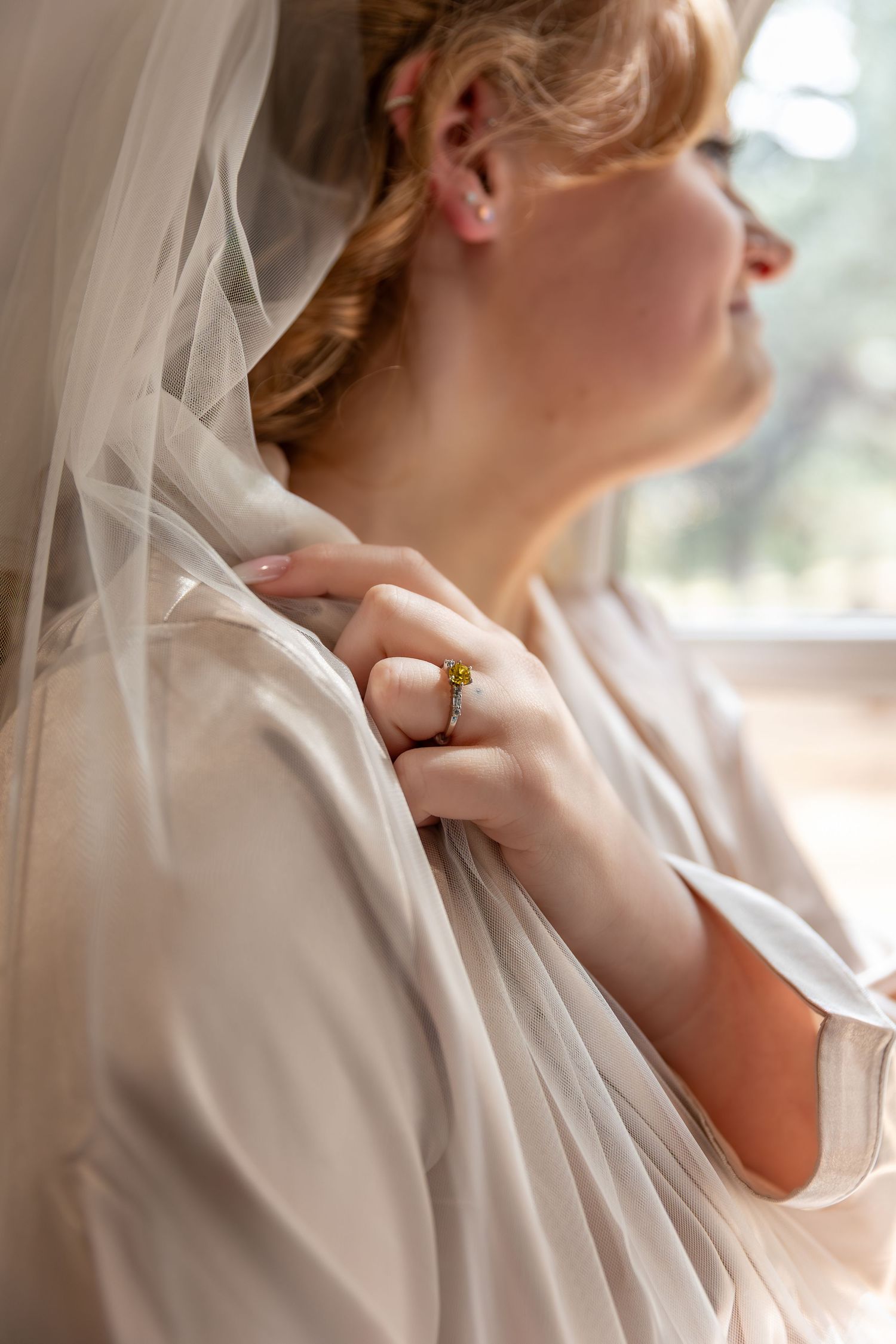 Bride in white robe and veil holds fabric, wearing gold ring, gazing out window on wedding day.