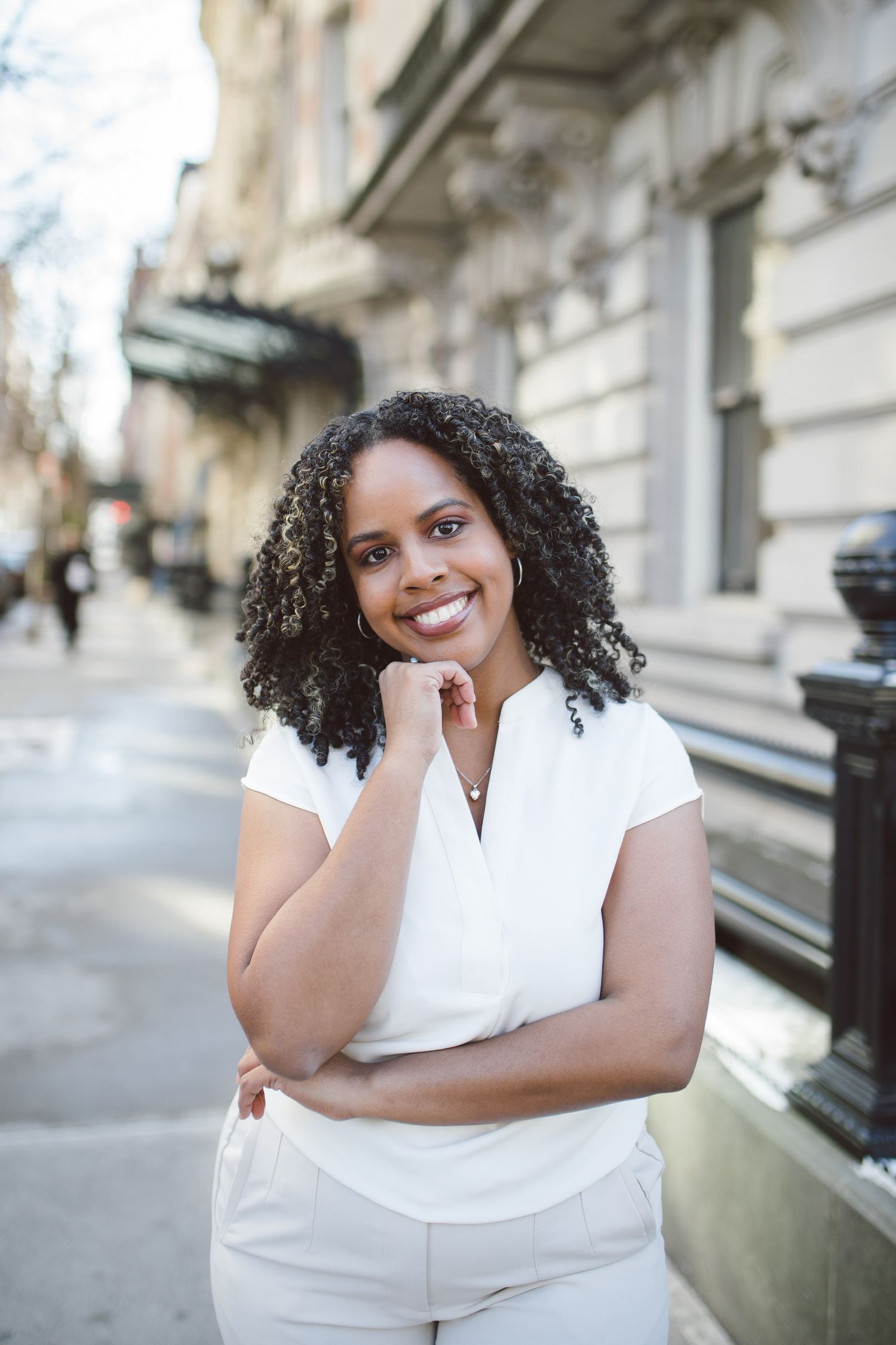 Professional in a crisp white outfit poses confidently on a city street with natural curly hair and warm smile.