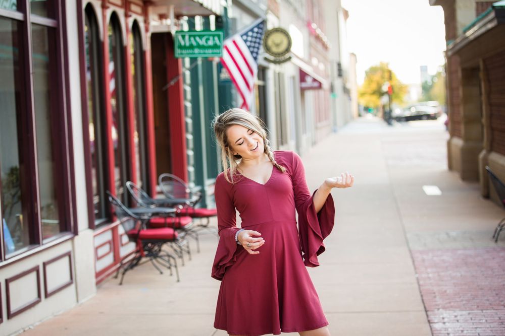 Person in burgundy dress twirling on a small-town sidewalk with American flag and storefronts visible.