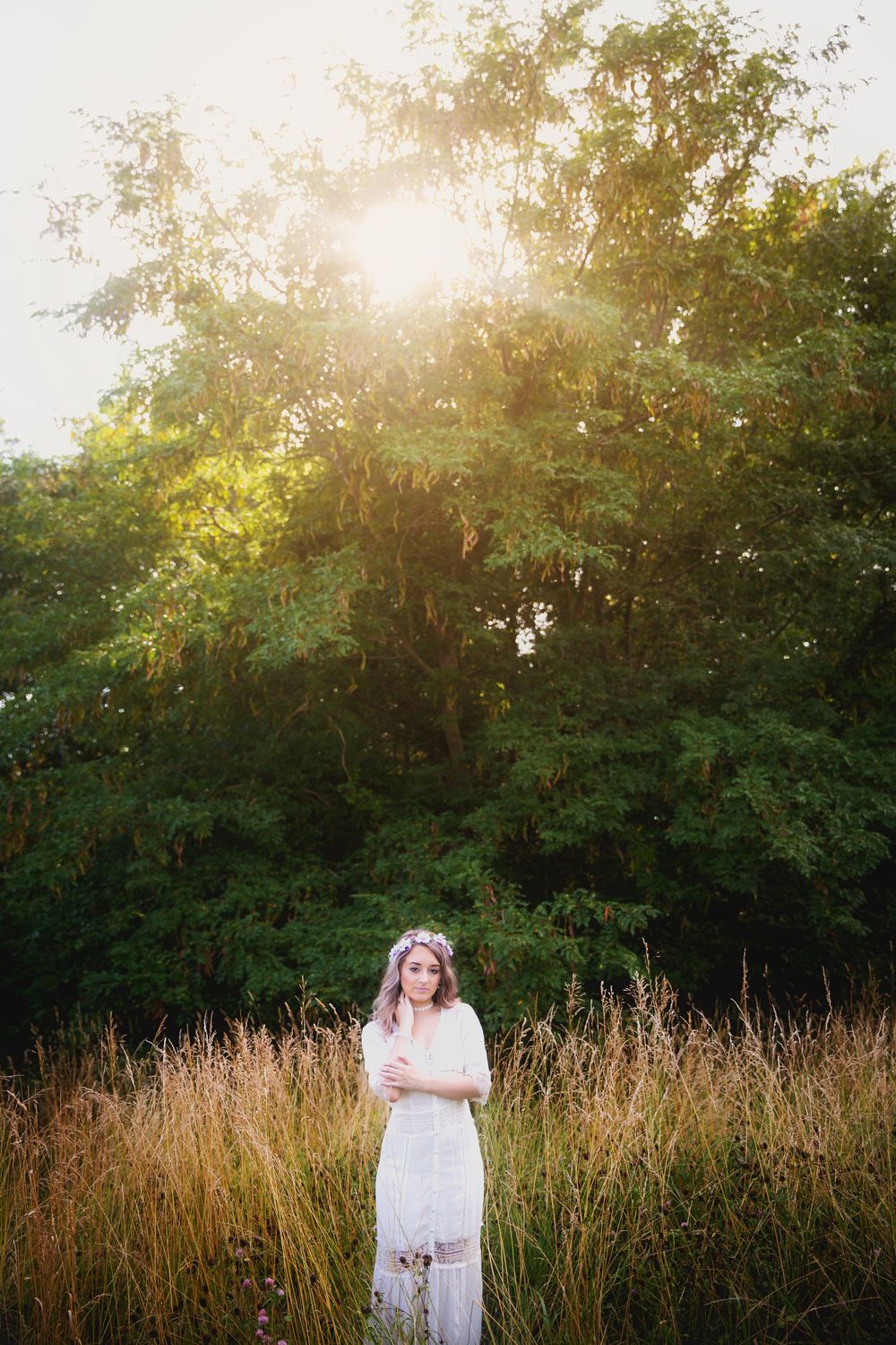A figure in a white dress standing in tall golden grass during sunset with trees in background.
