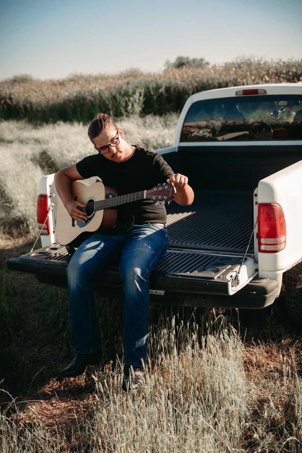 Musician playing acoustic guitar while sitting on pickup truck bed in rural field at sunset.