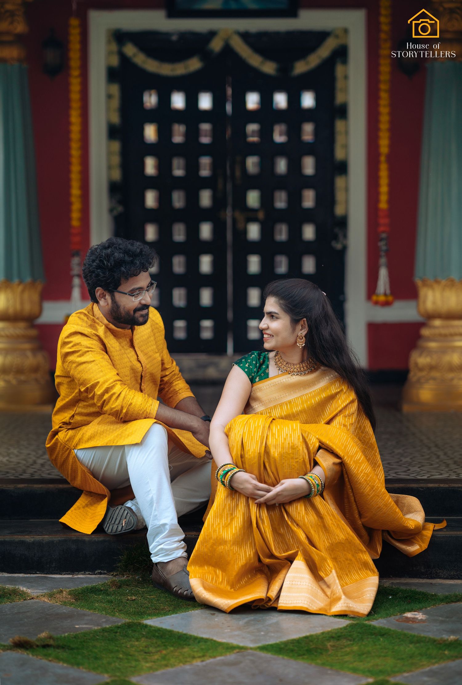 Couple in matching yellow traditional Indian outfits sitting together in front of temple doors.