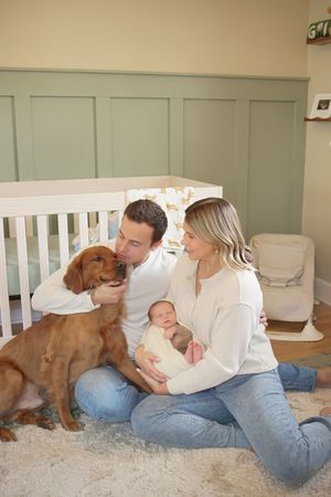 Young parents sit on a cozy nursery rug holding their newborn baby while kissing their golden retriever beside a white crib.
