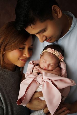 Smiling mother and father gently cradle their sleeping newborn daughter wearing a pink wrap and floral headband.
