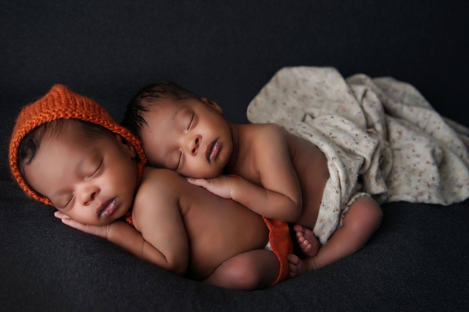 Two sleeping newborn twins nestle together on a dark blanket, one in an orange knit bonnet.