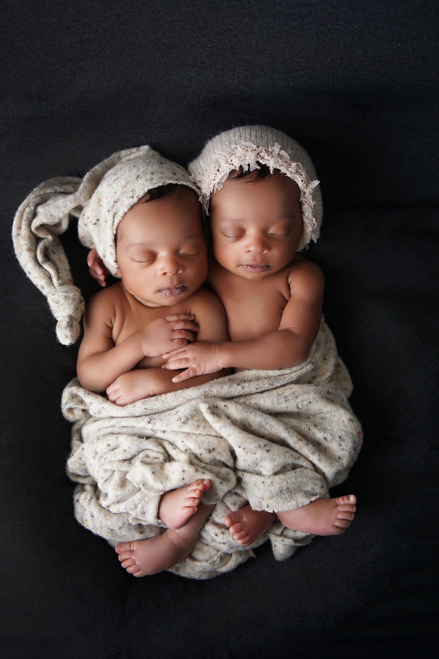 Newborn twins in cream knit hats sleep cuddled together, wrapped in a soft speckled blanket.
