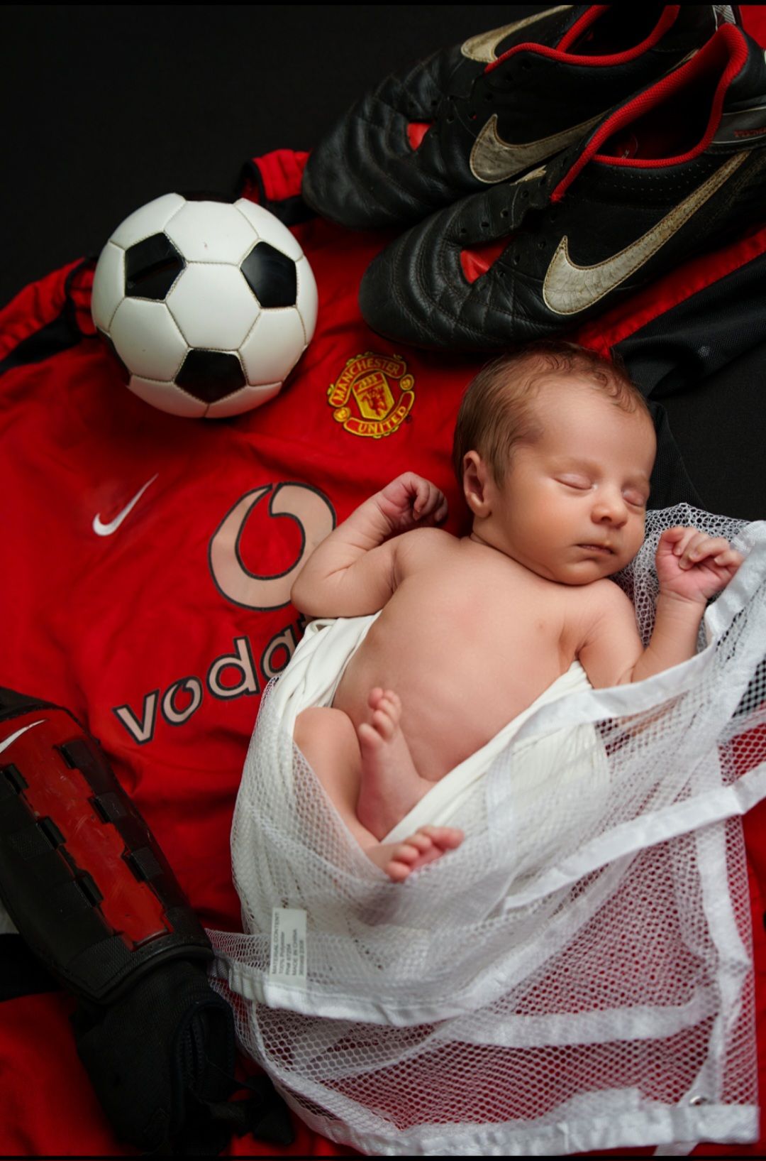 Sleeping newborn wrapped in a mesh bag beside a soccer ball, cleats and a red Manchester United jersey.