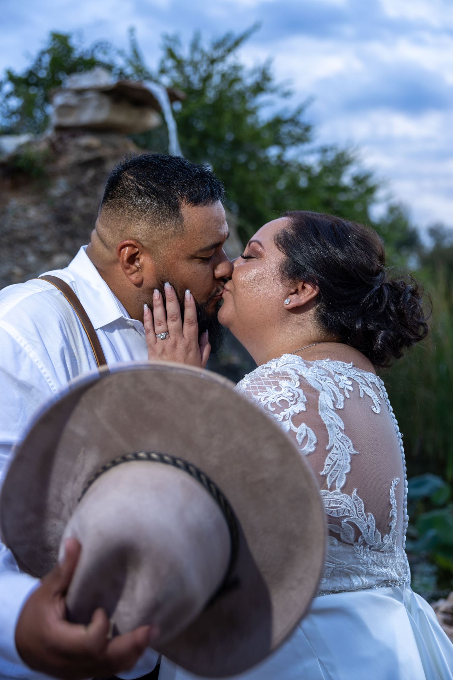 A romantic wedding portrait with a brown cowboy hat in the foreground against a natural backdrop.