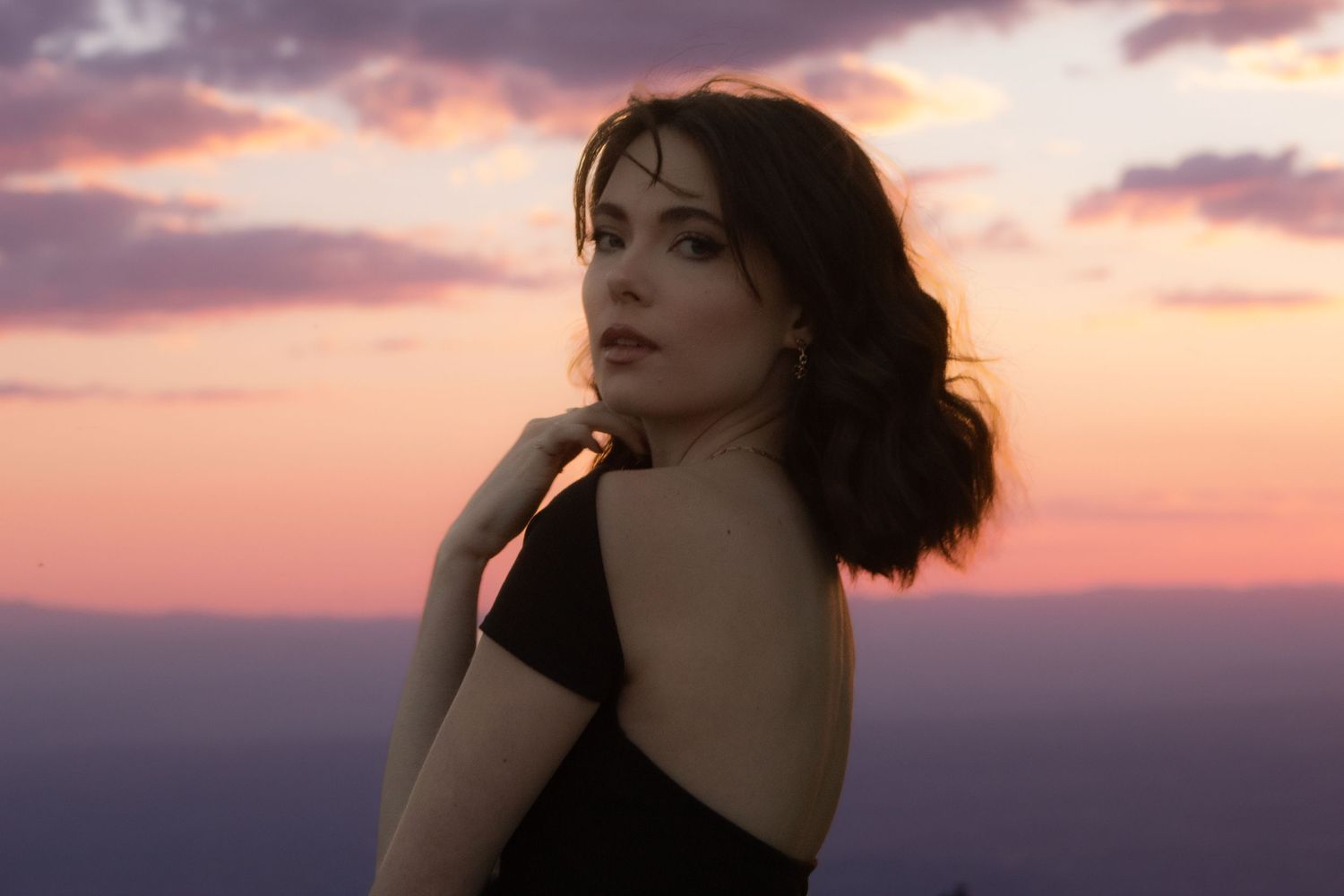 Woman in black dress poses against dramatic sunset sky with pink and purple clouds.