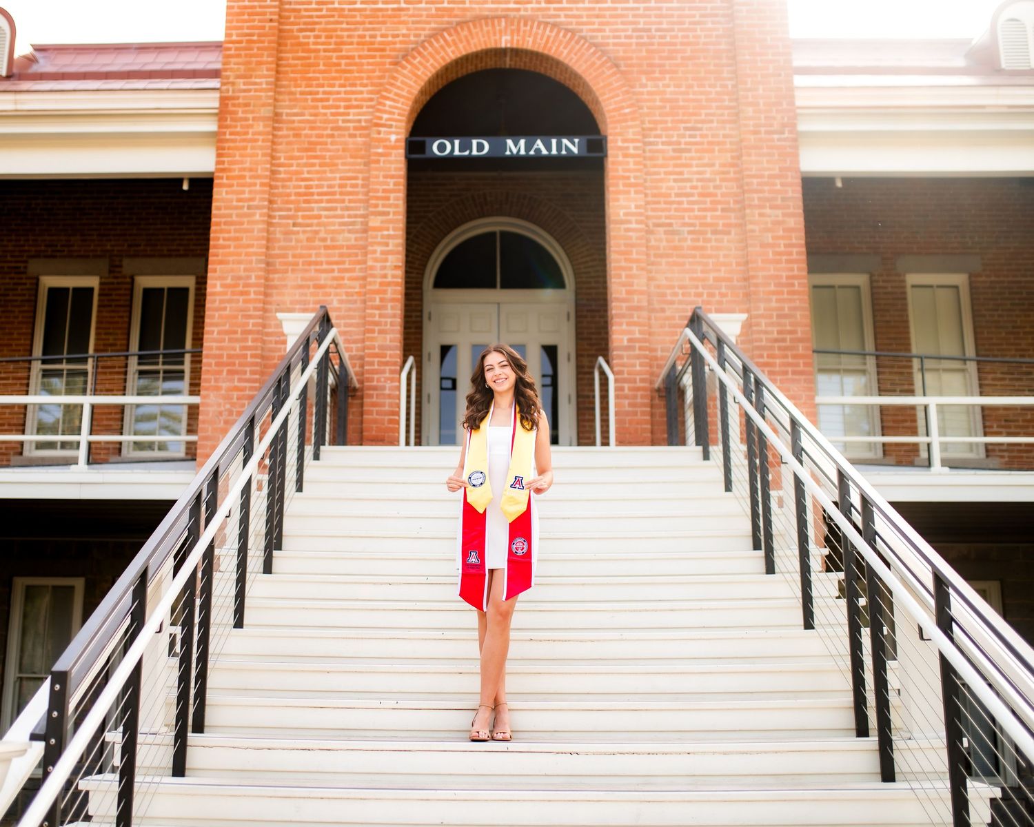 Graduate in red and white sash standing on steps outside historic Old Main university building.