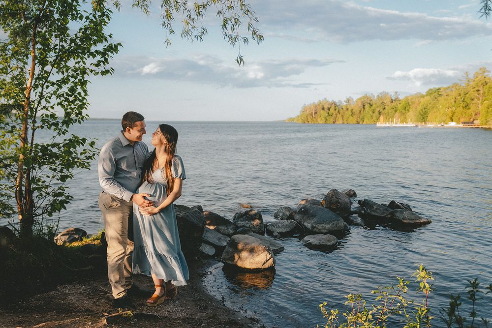 Couple embracing by rocky lakeside shore surrounded by lush green trees and calm blue water at golden hour.