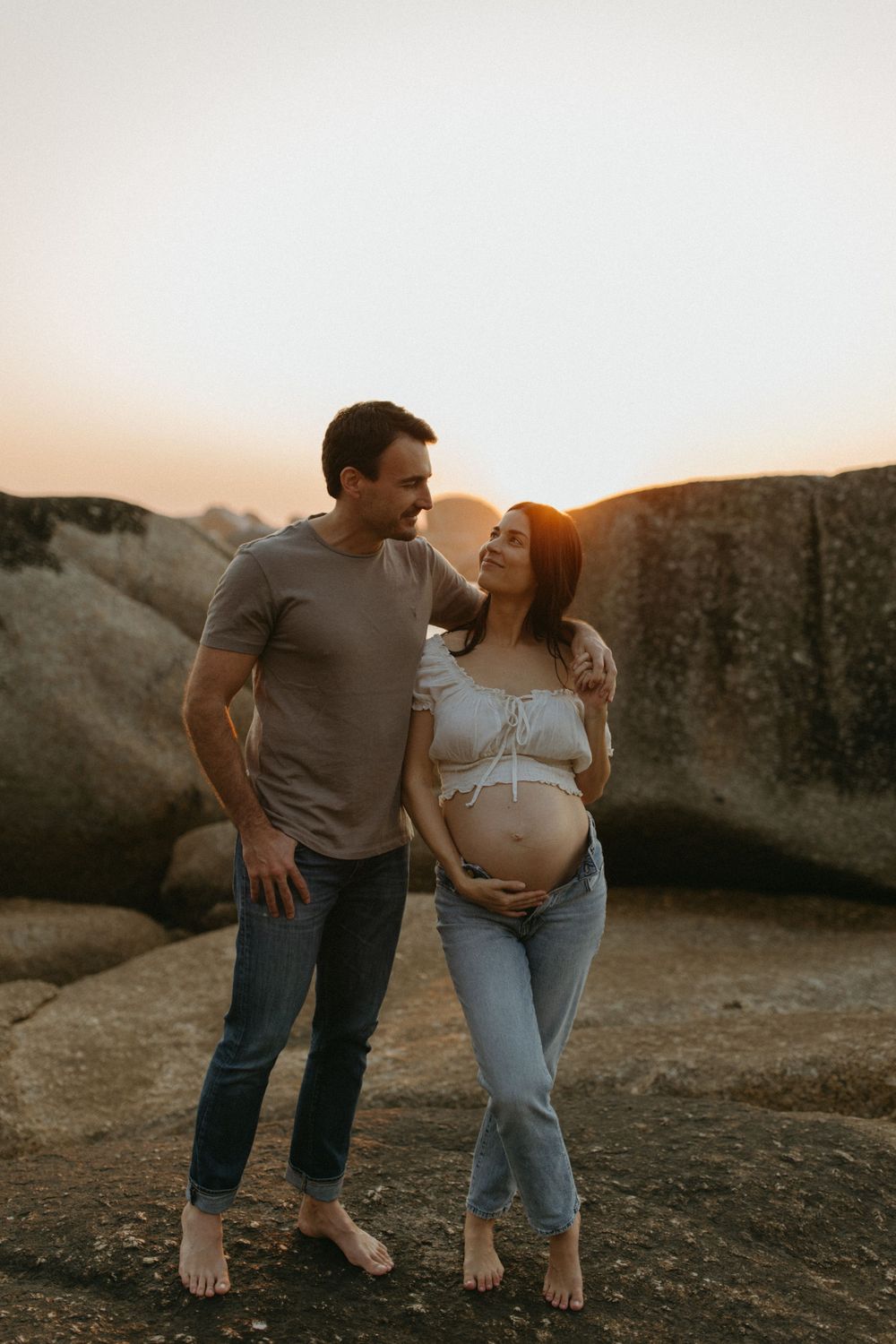 Couple shares intimate moment during sunset maternity photoshoot on rocky terrain.