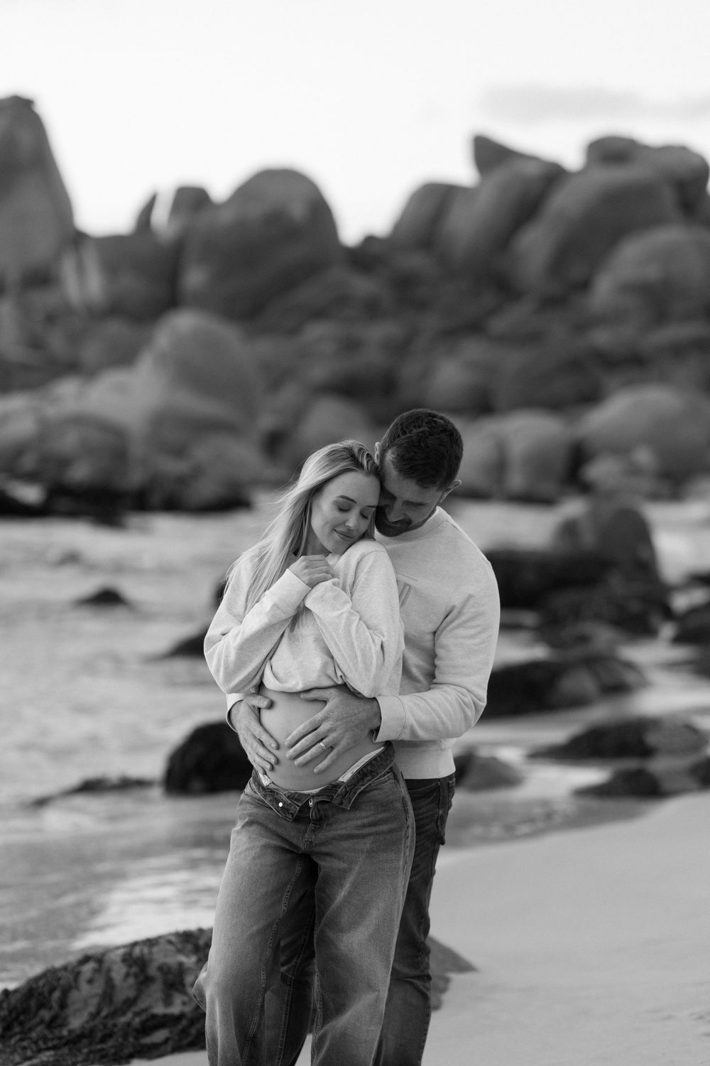 A romantic black and white photo of a couple embracing on a rocky beach with boulders in the background.