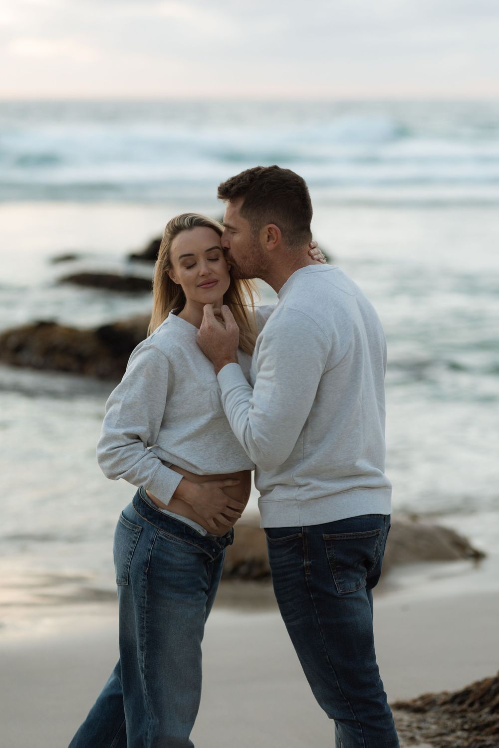 A couple in matching white sweaters and jeans share an intimate moment on a rocky beach at sunset.