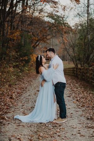 A couple poses for maternity photos on a wooded path covered in fall foliage.