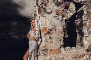 Woman in white clothing poses against rustic stone wall backdrop.