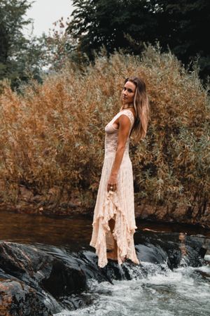 Model in flowing white dress stands on rocks near rushing stream.