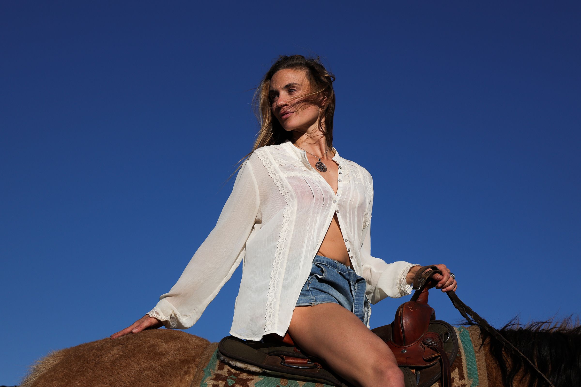Model poses on horseback wearing white shirt and denim shorts against bright blue sky.