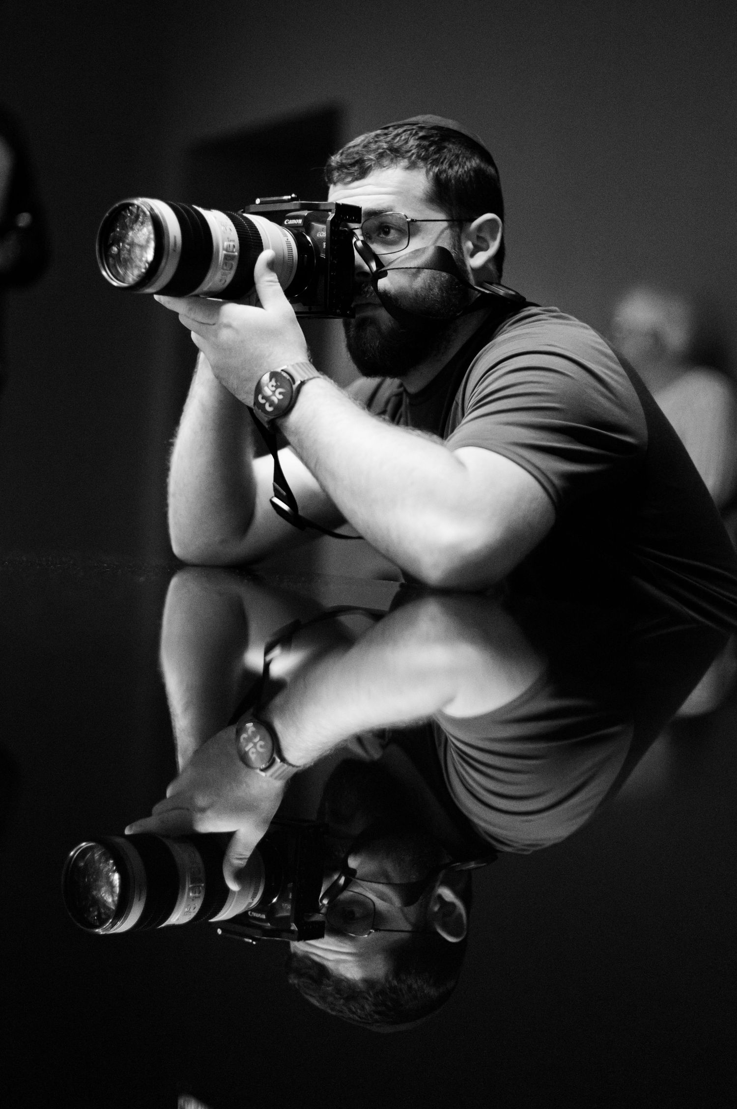 Bearded male photographer crouches with camera and telephoto lens in black and white portrait.