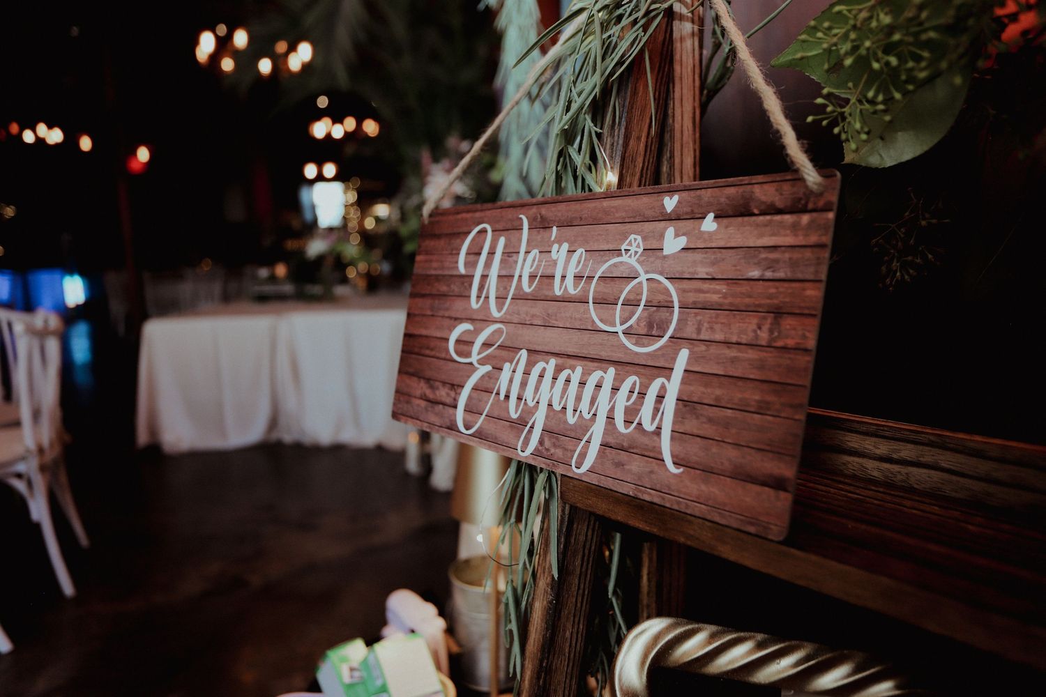 Rustic wooden We're Engaged sign hanging at an outdoor engagement party with string lights and greenery.