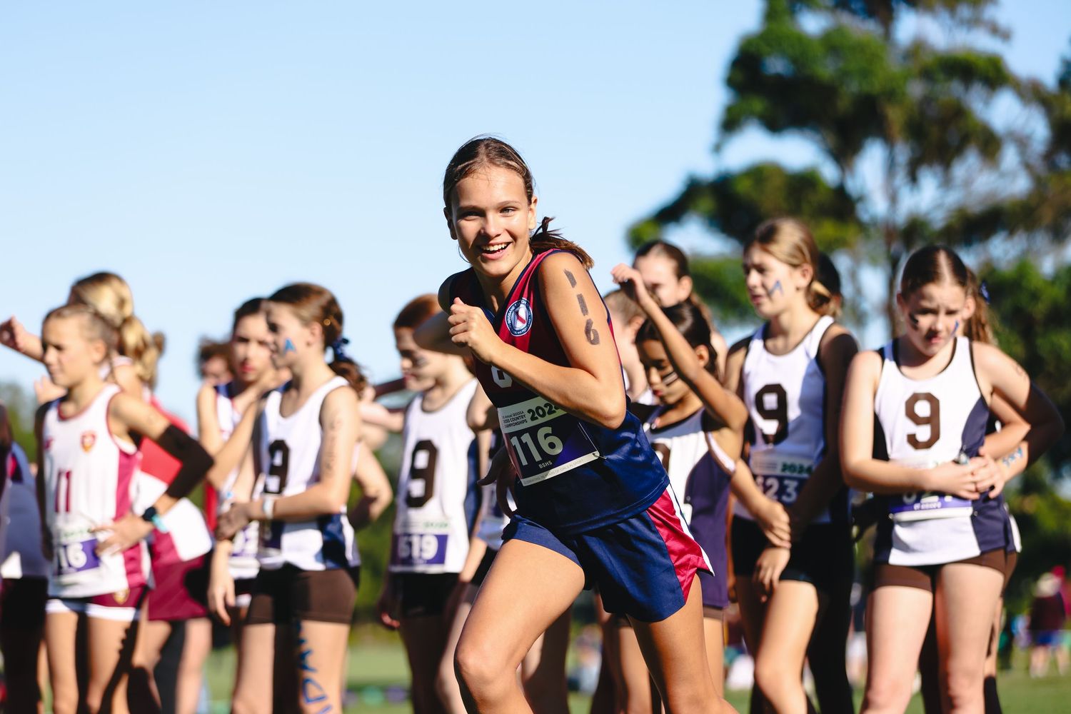 Athletes running in a competitive cross country race wearing numbered bibs on sunny day.
