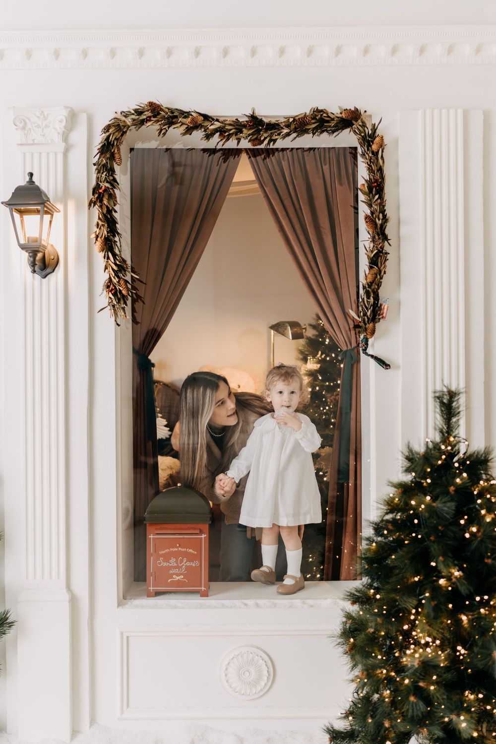 A cozy Christmas window scene with brown curtains, white columns, and decorated trees glowing with warm lights.